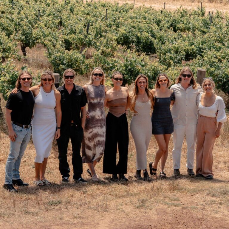 A group of people smiling, in front of a row of grapevines enjoying the Waves of Wine Premium Margaret River Wine Tour